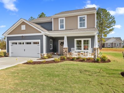 A two-story craftsman-style home with gray and beige siding, white trim, and stone accents on the front porch columns. The house features a two-car garage, covered front porch with white rocking chairs, and professional landscaping with mulched beds and a manicured lawn. The architecture includes craftsman details like white pillars, divided light windows, and board and batten siding accents on the upper level.