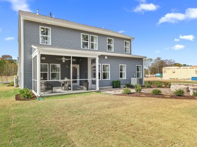 A rear view of a two-story home featuring blue-gray siding with white trim. The back of the house includes a covered porch with screening and outdoor furniture, large windows throughout, and a professionally landscaped yard with mulched beds and stone retaining walls. In the background, new construction can be seen in the developing neighborhood. The home is positioned on a gently sloping lawn with young landscaping.