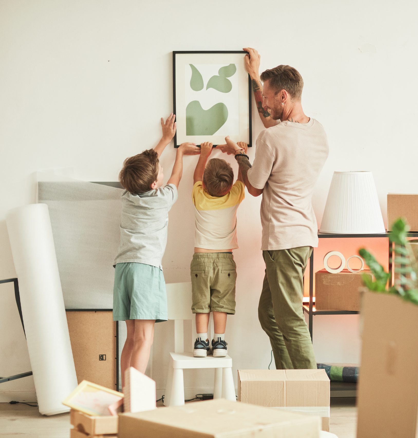 Full length portrait of happy father with two sons hanging pictures on wall while moving in to new home